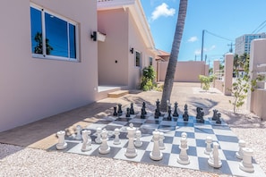 A chessboard set up on a patio with a palm tree and buildings in the background. - Summer Fun · 6 King BR Pool Villa, Sleeps 14 · 5-Min Walk to Palm Beach (Noord)