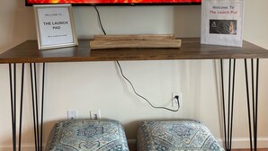 A TV on a wooden TV stand displaying a fiery landscape, a welcome sign, and two patterned ottomans. - The Launch Pad - Perfect for viewing launches - minutes from beaches (Titusville)