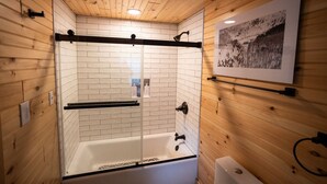 Een badkamer met houten lambrisering, een douche met glazen wand, witte betegelde muren en een ingelijste foto aan de muur.