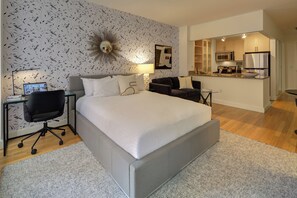 A modern hotel room with a large bed, a desk with a computer, a chair, and a sofa. There is a kitchen area visible in the background. - Bright Downtown Apartment | Doorman & Lounge (New York)