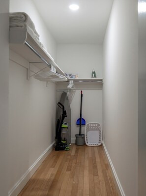 A narrow hallway with a wooden floor, a shelf with folded towels, a vacuum cleaner, a mop, and a laundry basket. - Bright Downtown Apartment | Doorman & Lounge (New York)