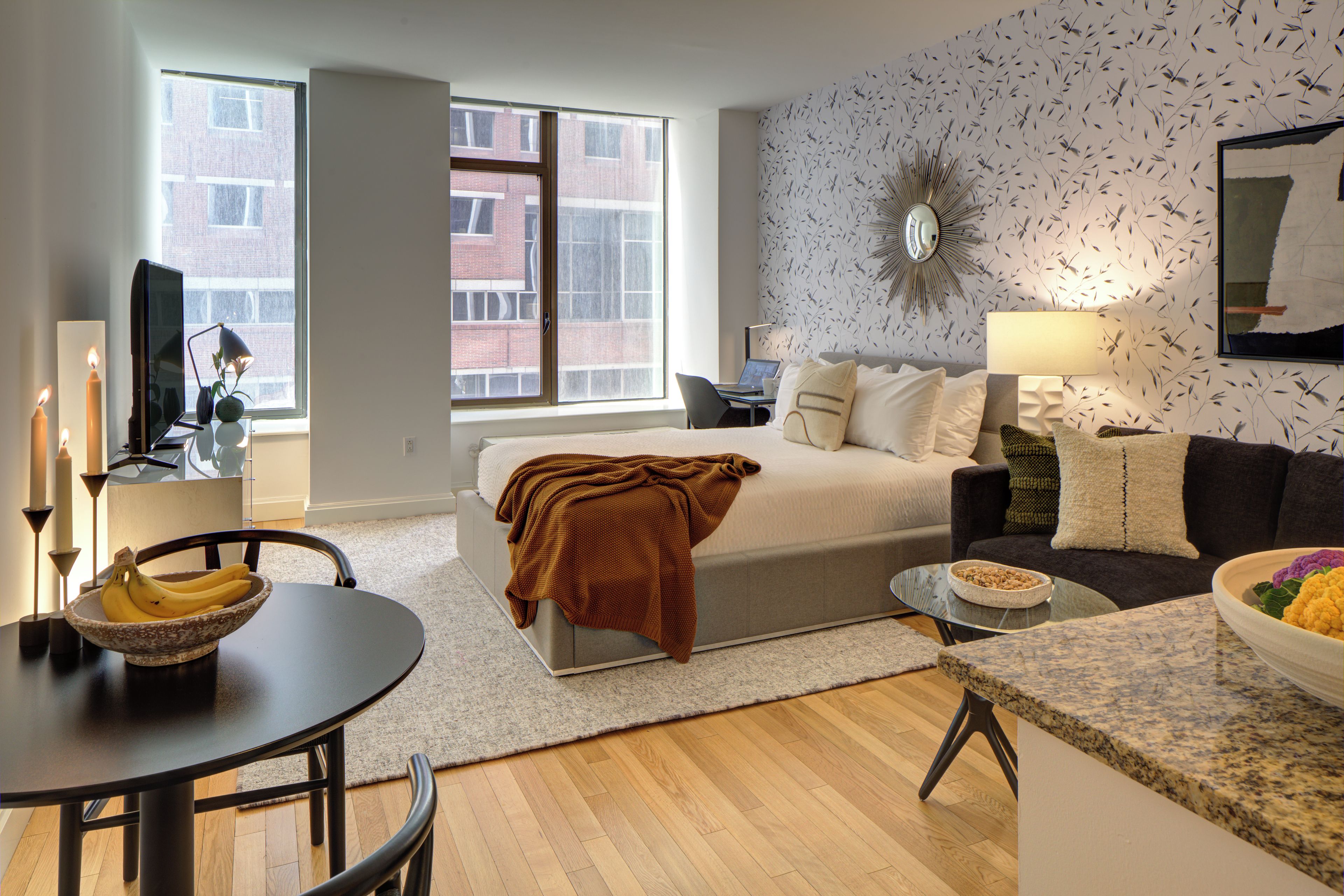 A modern living room with a bed, a sofa, a dining table with a bowl of fruit, and a TV.