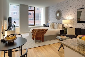 A modern living room with a bed, a sofa, a dining table with a bowl of fruit, and a TV. - Bright Downtown Apartment | Doorman & Lounge (New York)