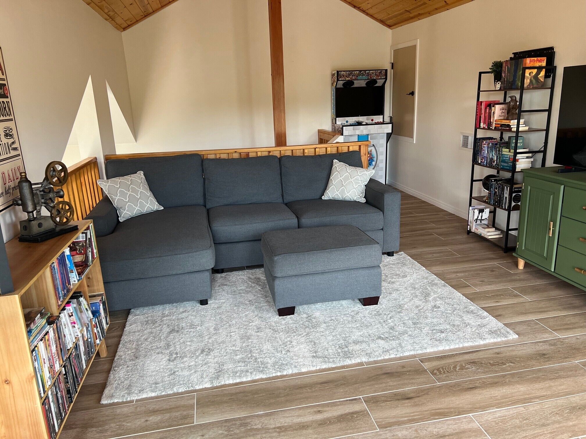 Ein Wohnzimmer mit einem grauen Ecksofa, einem kleinen Hocker, einem Holztisch, einem Bücherregal und einem grünen Schrank.