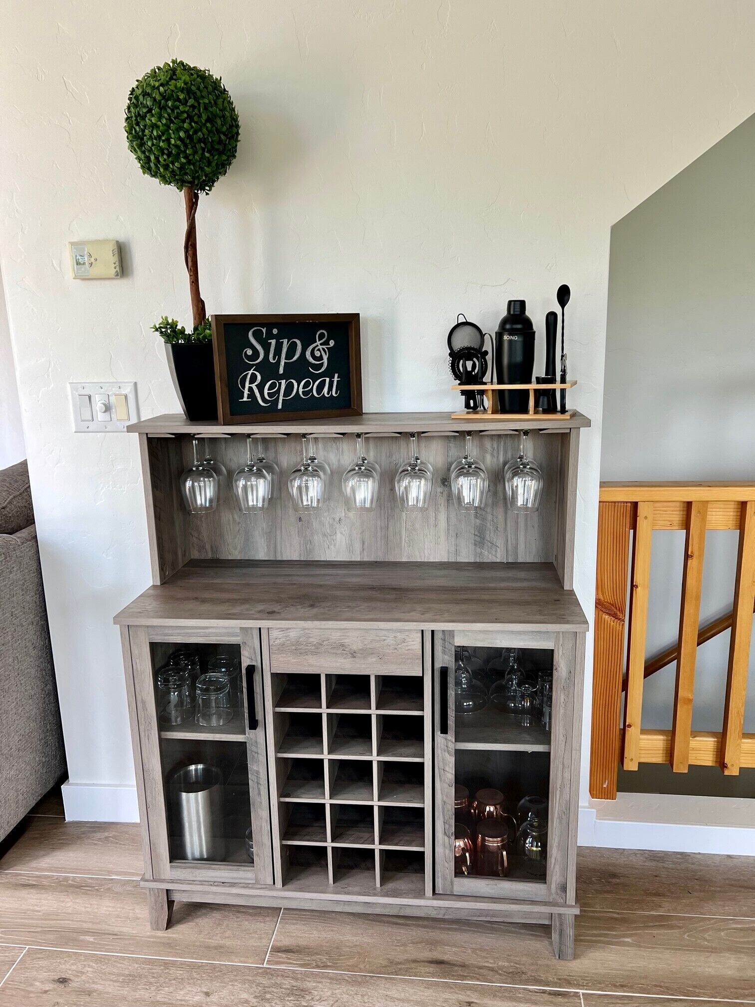 A wooden bar area with a sign that reads "Sip & Repeat," a potted plant, and a shelf with black bottles and a spoon.
