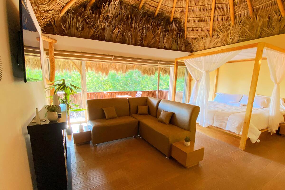 A room with a thatched roof, a sofa, a bed with a canopy, and a view of greenery outside.