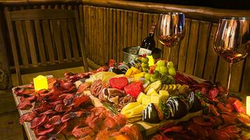A table set with a variety of fruits, cheeses, and a bottle of wine, placed on a wooden deck with a city view in the background.