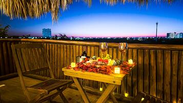 An outdoor dining setup with a table set for two, featuring a bottle of wine, glasses, and a variety of fruits and chocolates.