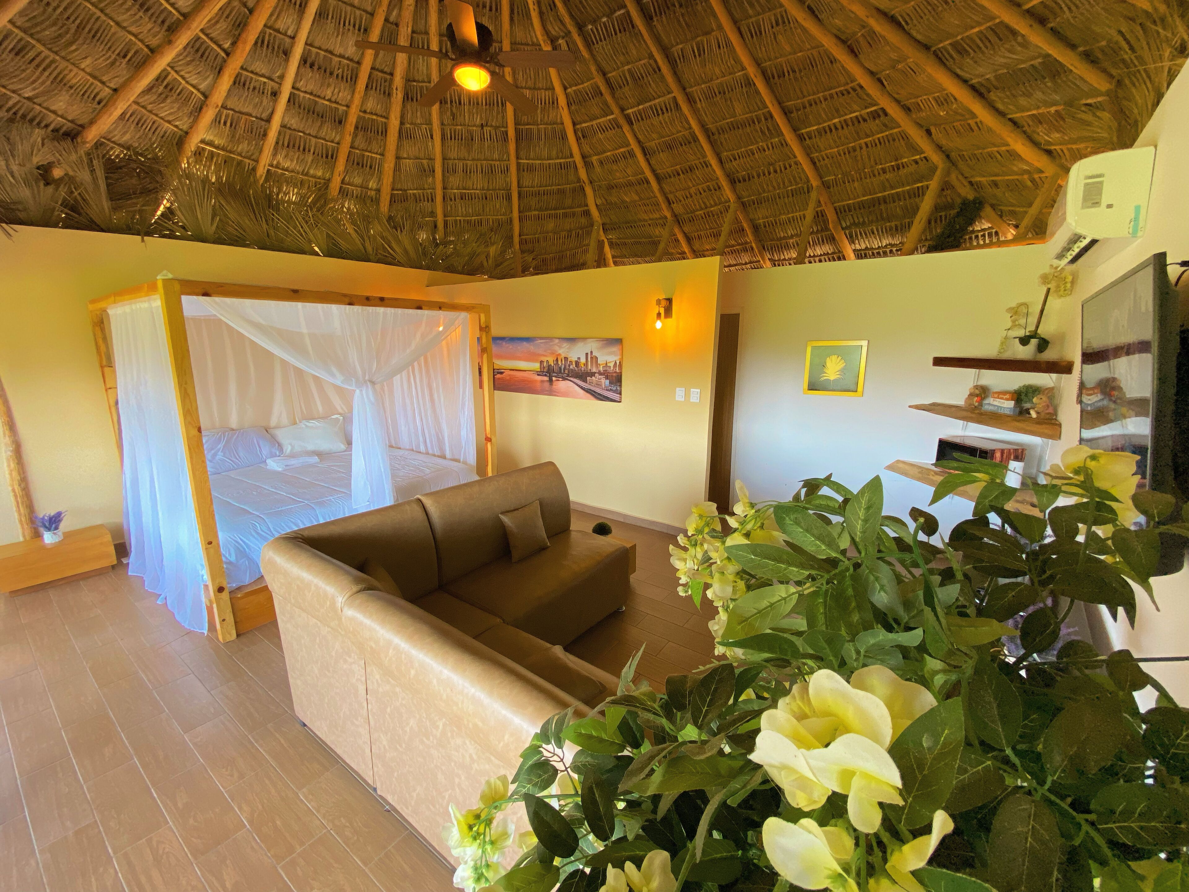 Una habitación con una cama bajo un tejado de paja, un sofá y una planta grande.