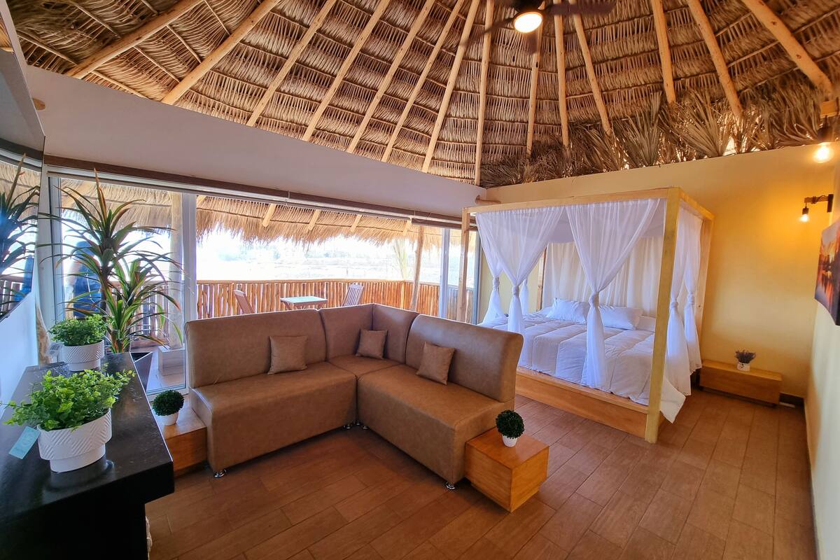 A room with a thatched roof, a sofa, a bed with a canopy, and potted plants.