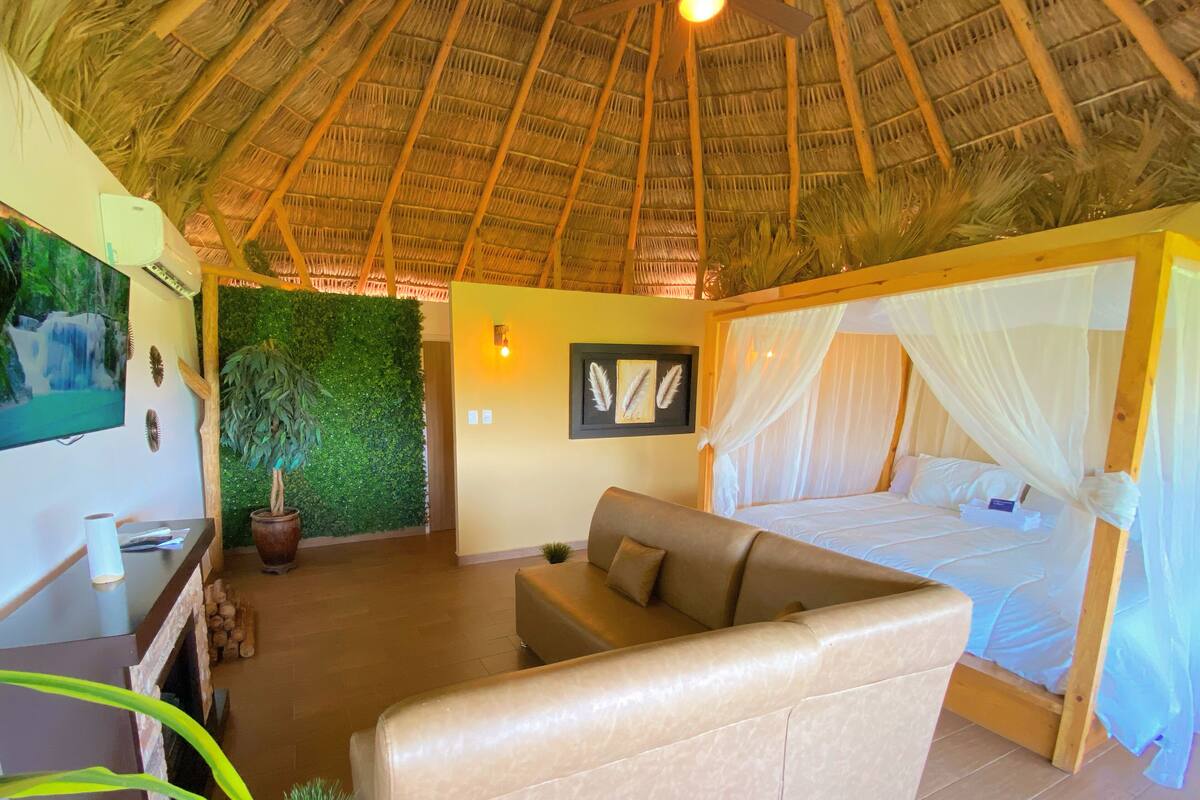 A room with a thatched roof, a bed with a canopy, a sofa, a desk with a plant, and a green wall.