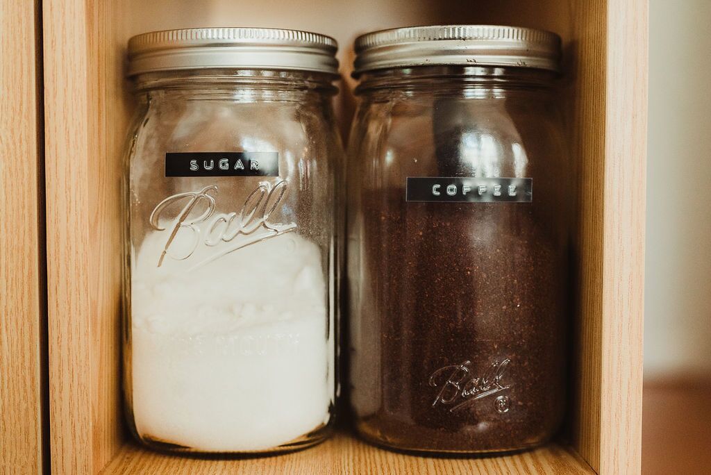 Deux bocaux en verre, munis de couvercles en métal, dont l’un est étiqueté « SUCRE » et l’autre « CAFÉ », sont placés dans un placard en bois.