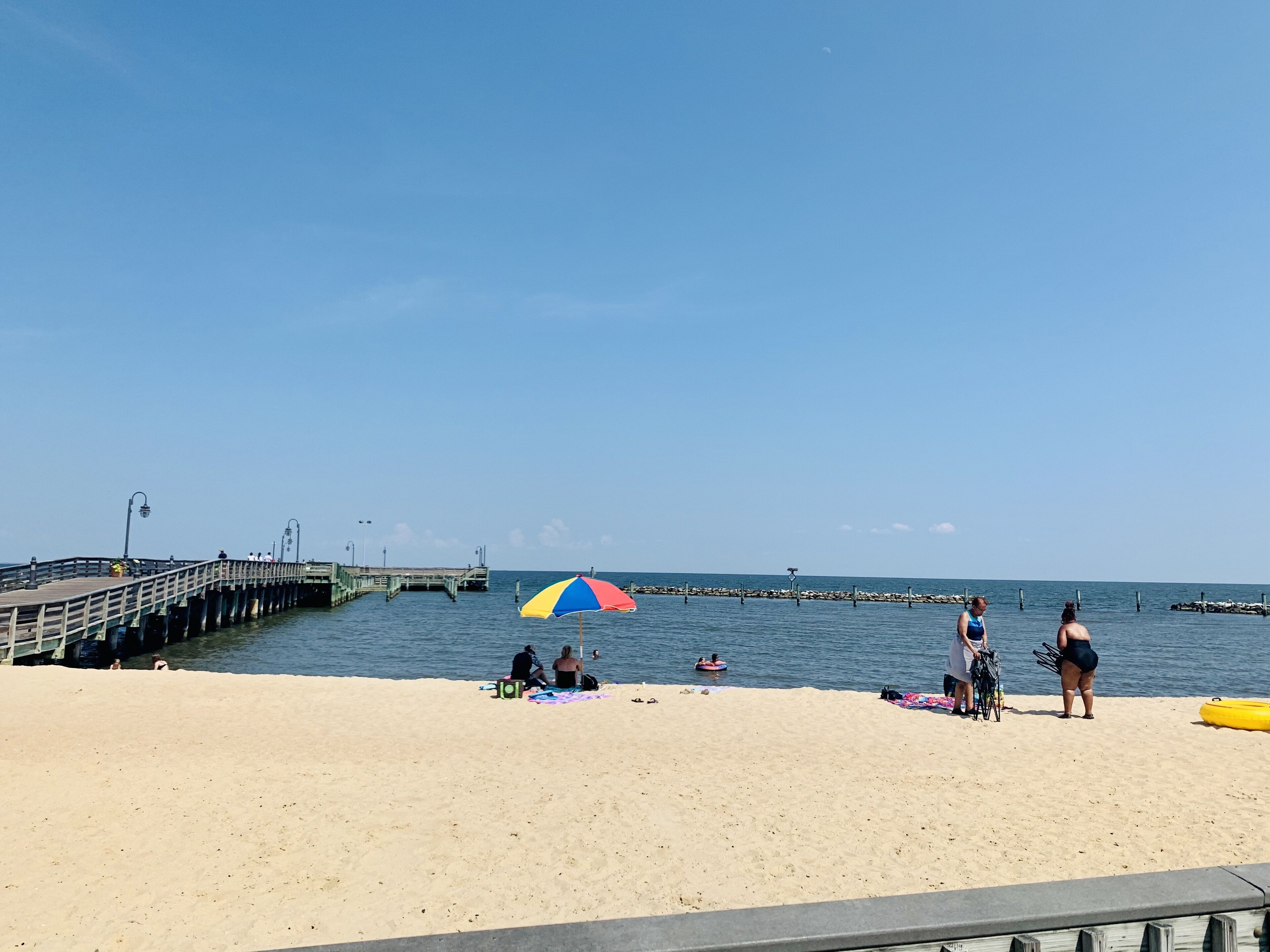 Een strand met een steiger die het water in reikt, mensen ontspannen op het zand, en een kleurrijke parasol die schaduw biedt.