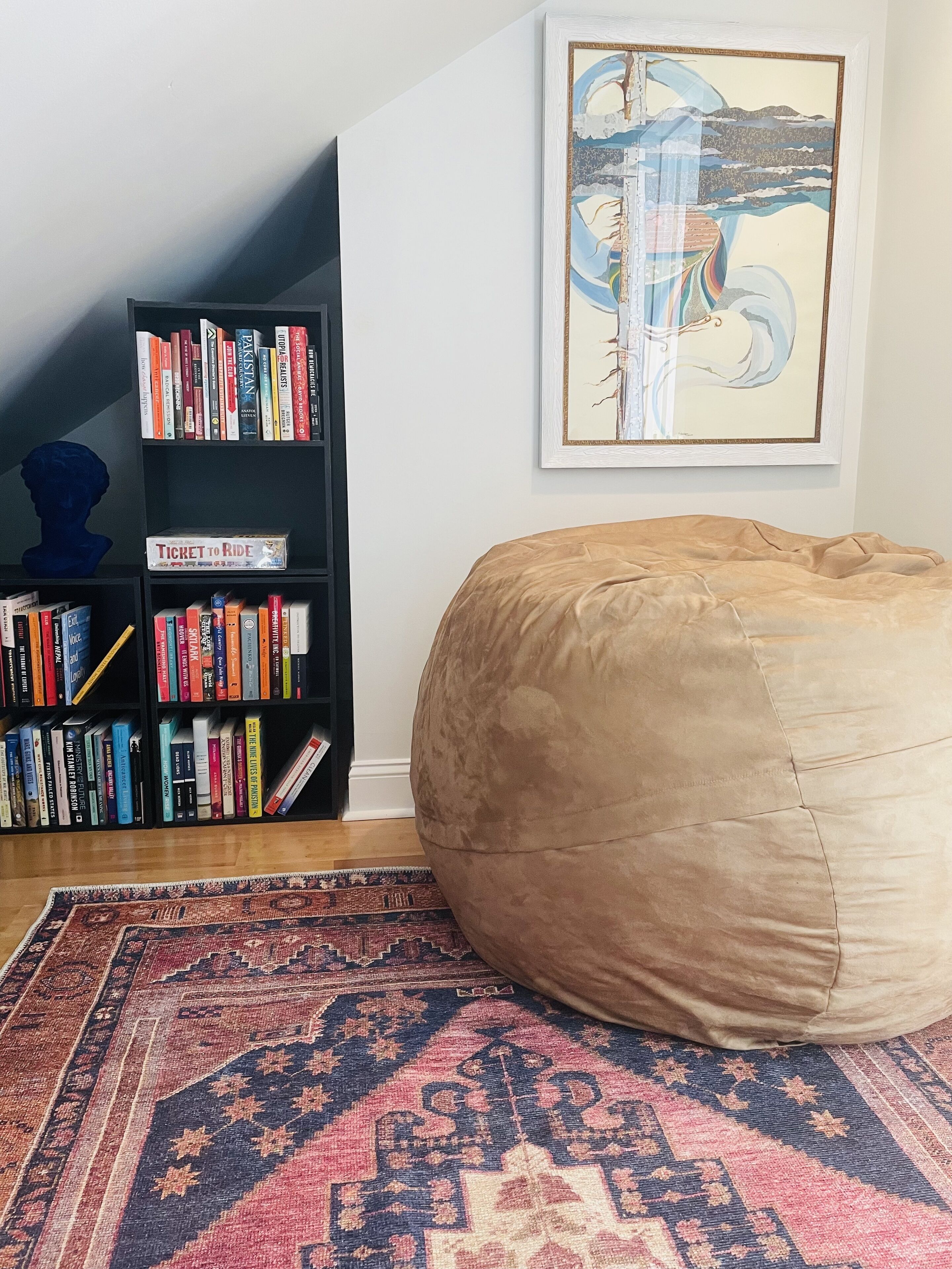 Una habitación con un sillón informal color beige, una estantería llena de libros, una obra de arte enmarcada en la pared y una alfombra estampada en el suelo.