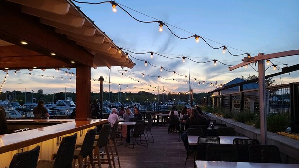 Un espacio de comedor al aire libre con luces colgantes, mesas y sillas de madera, con vista a un puerto deportivo con barcos.