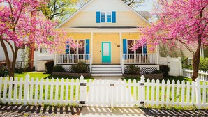 Una casa de dos pisos con un porche, cerca blanca de piquete y árboles en flor de color rosa.
