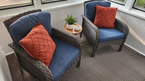 Une pièce avec deux fauteuils en osier, chacun agrémenté d’un coussin bleu et d’un oreiller orange. On y trouve une petite table en bois sur laquelle sont posés un pot de fleurs et un plateau de assiettes.