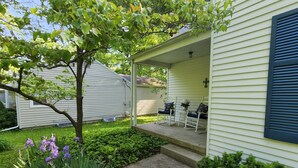Une maison avec un porche, un petit jardin avec des fleurs violettes et un arbre.