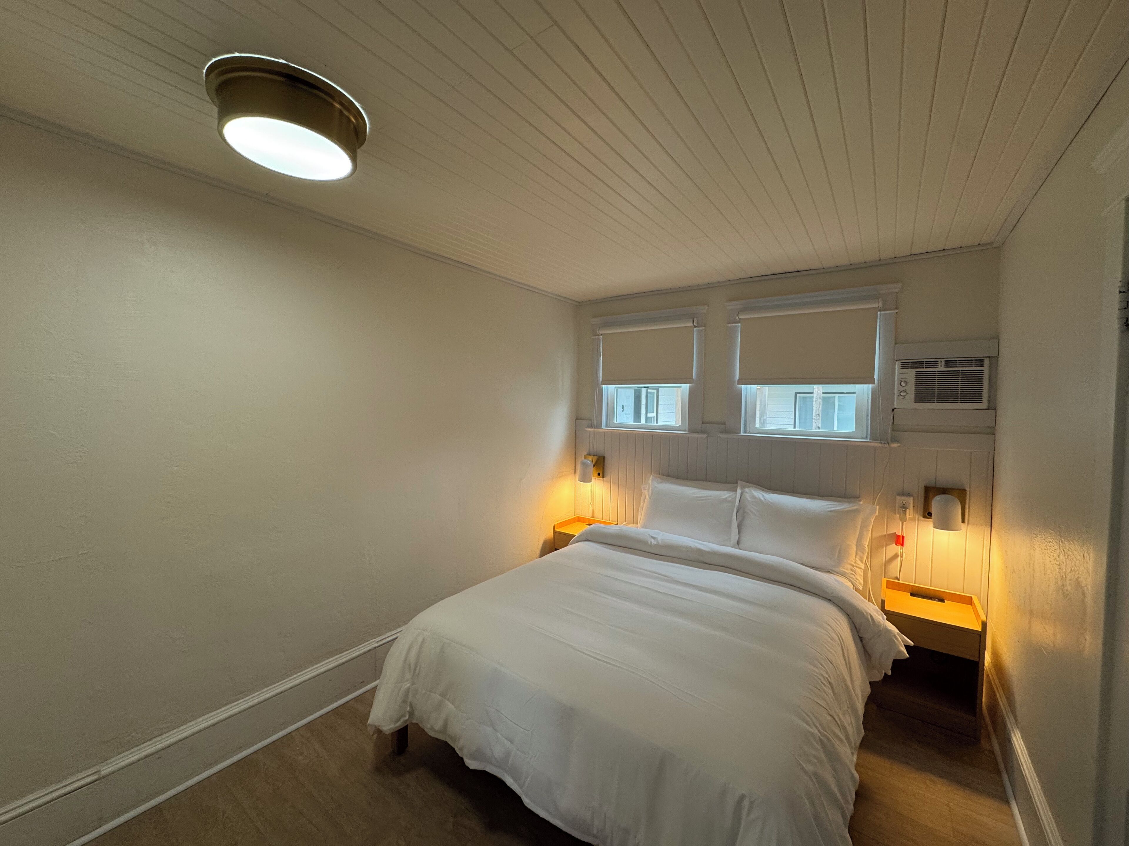 Un dormitorio con una cama, dos lámparas de noche, una lámpara de techo y una ventana con persianas.
