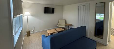 A room with a blue sofa, a small wooden table, a lamp, a TV on the wall, and a mirror.
