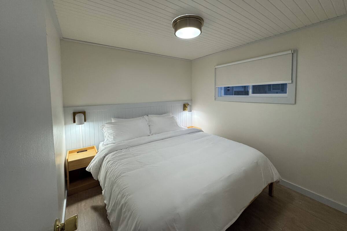 A bedroom with a single bed, a nightstand, a wall-mounted light, and a window with blinds.