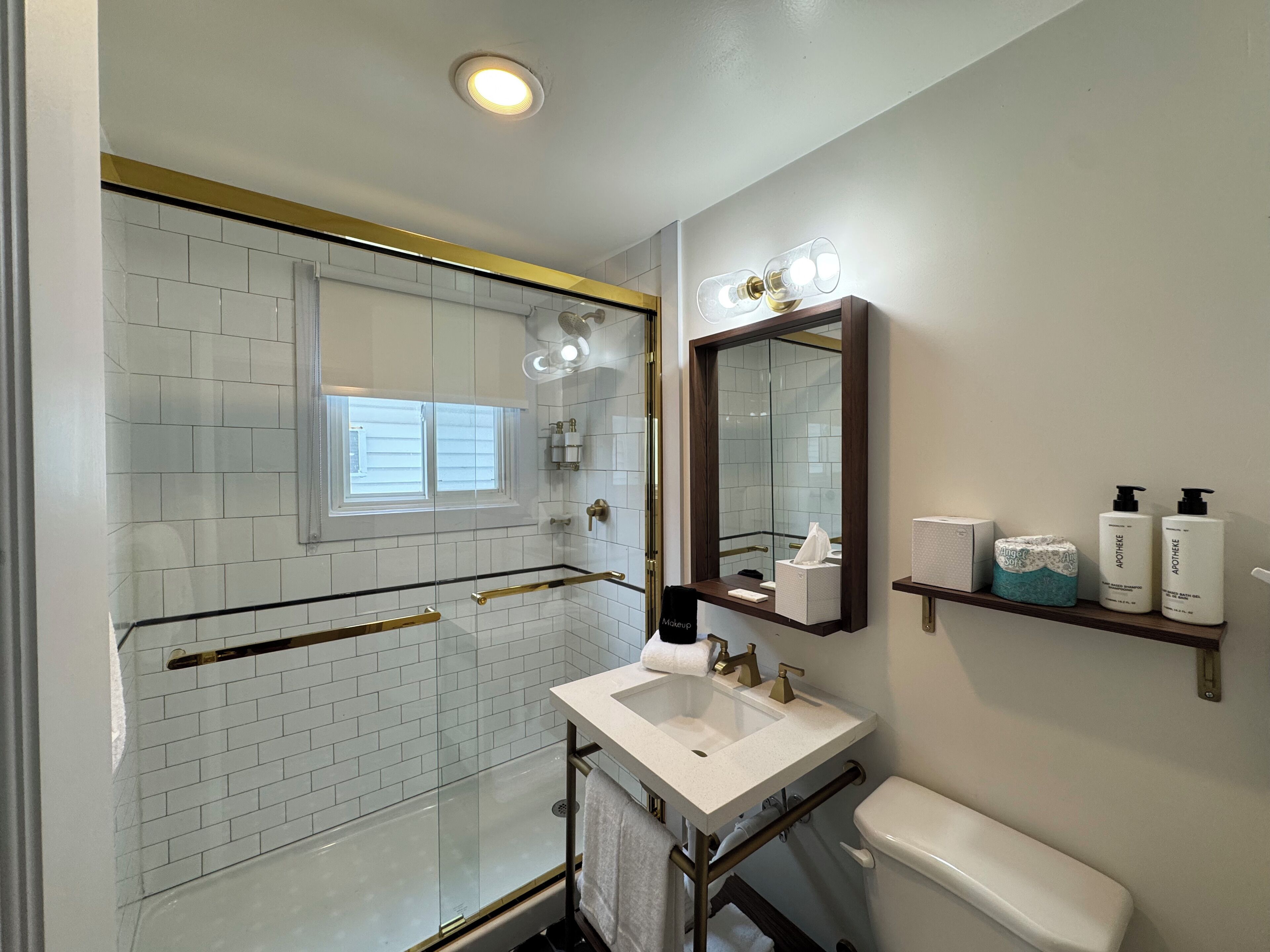 Un baño moderno con ducha de vidrio, lavamanos blanco y retrete. Hay estantes con artículos de aseo y un espejo grande sobre el lavamanos.