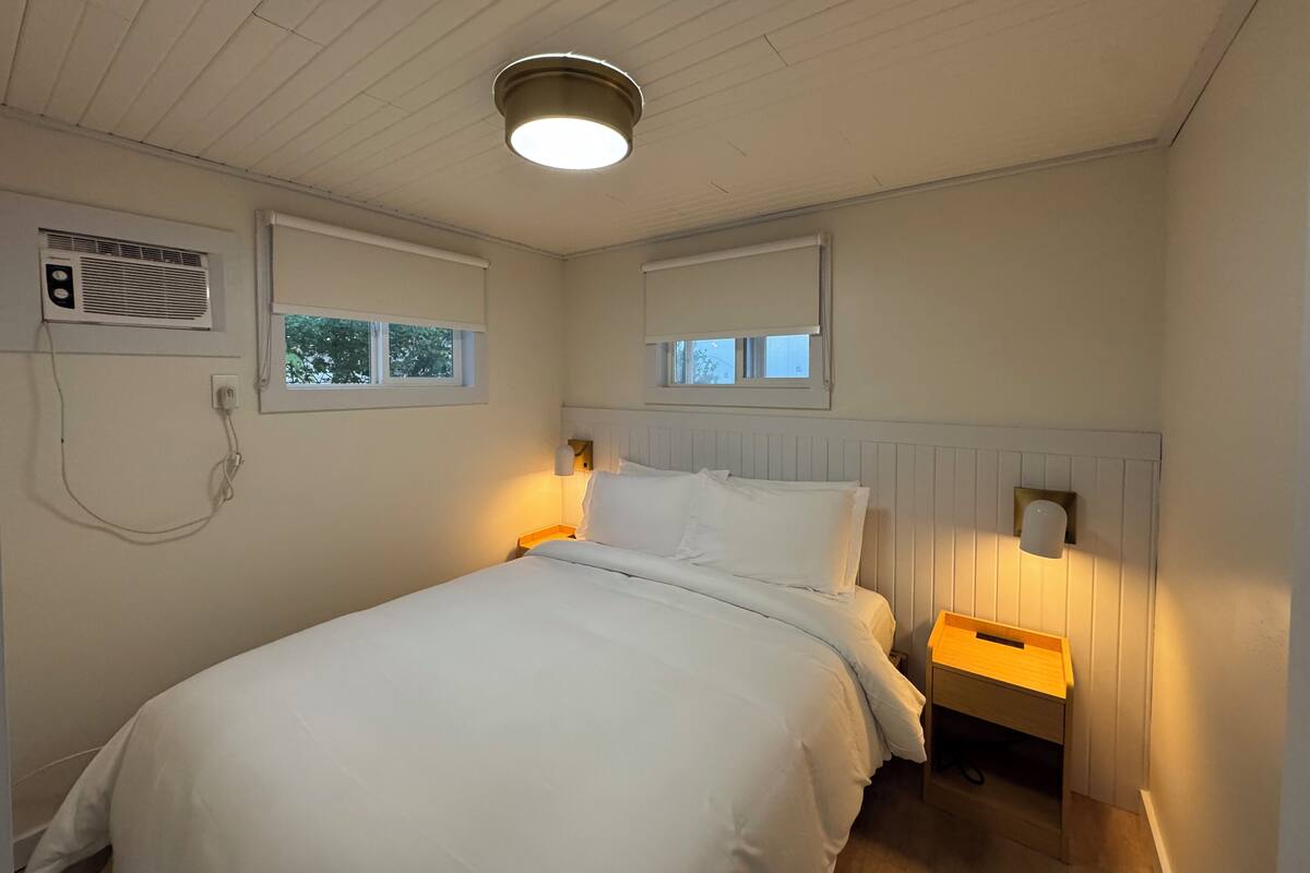 A bedroom with a bed, two bedside tables, a wall-mounted heater, and two windows with blinds.