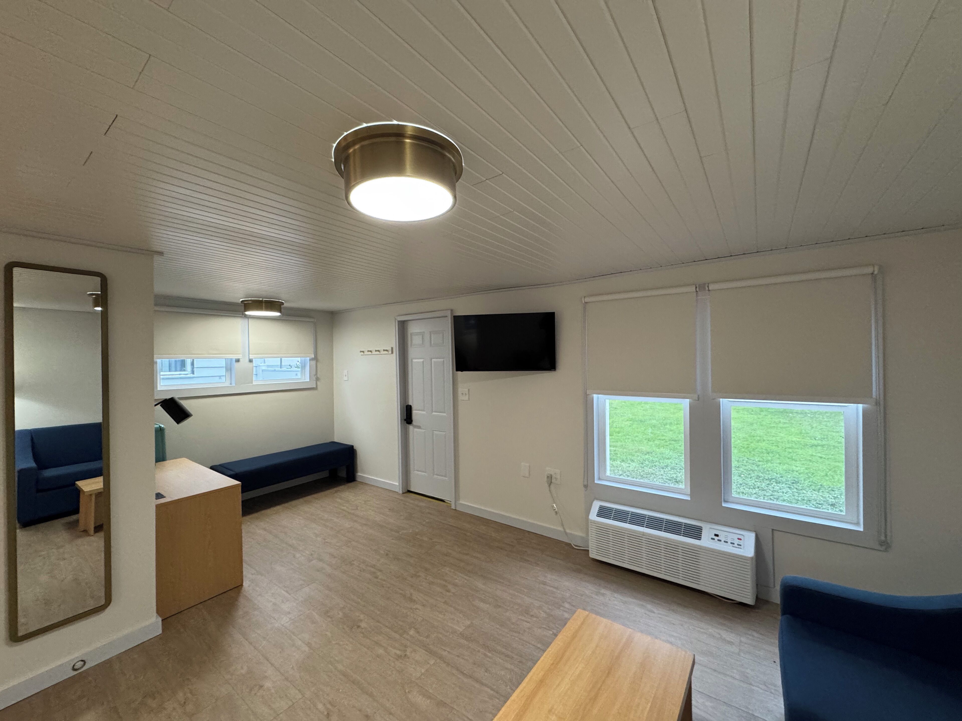 A room with a wooden floor, a blue sofa, a small table, a mirror, a ceiling light, a door, and a window with a view of the outdoors.
