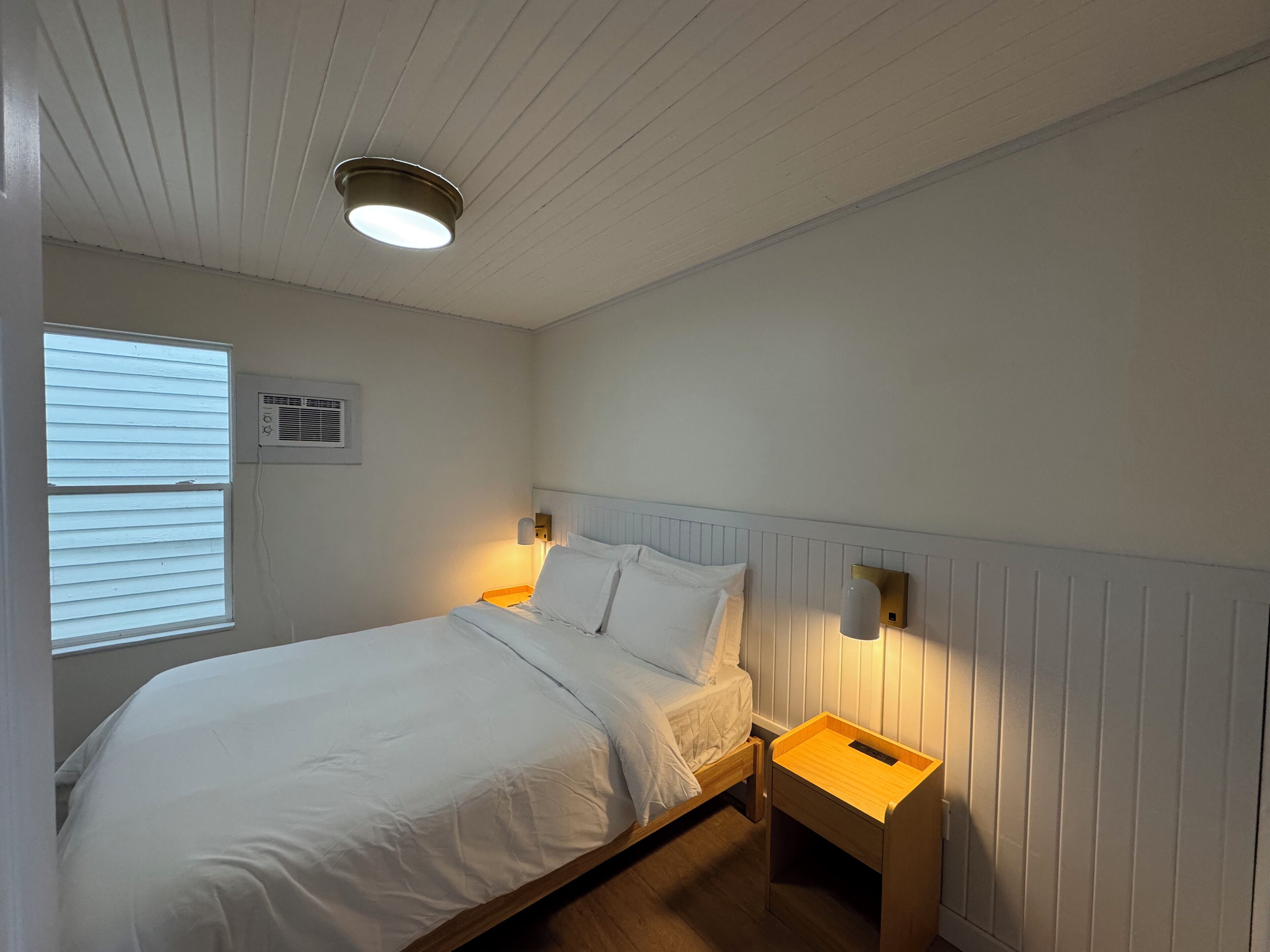 Un dormitorio con cama, mesitas de noche, luz empotrada en la pared, ventana con persianas y aire acondicionado.