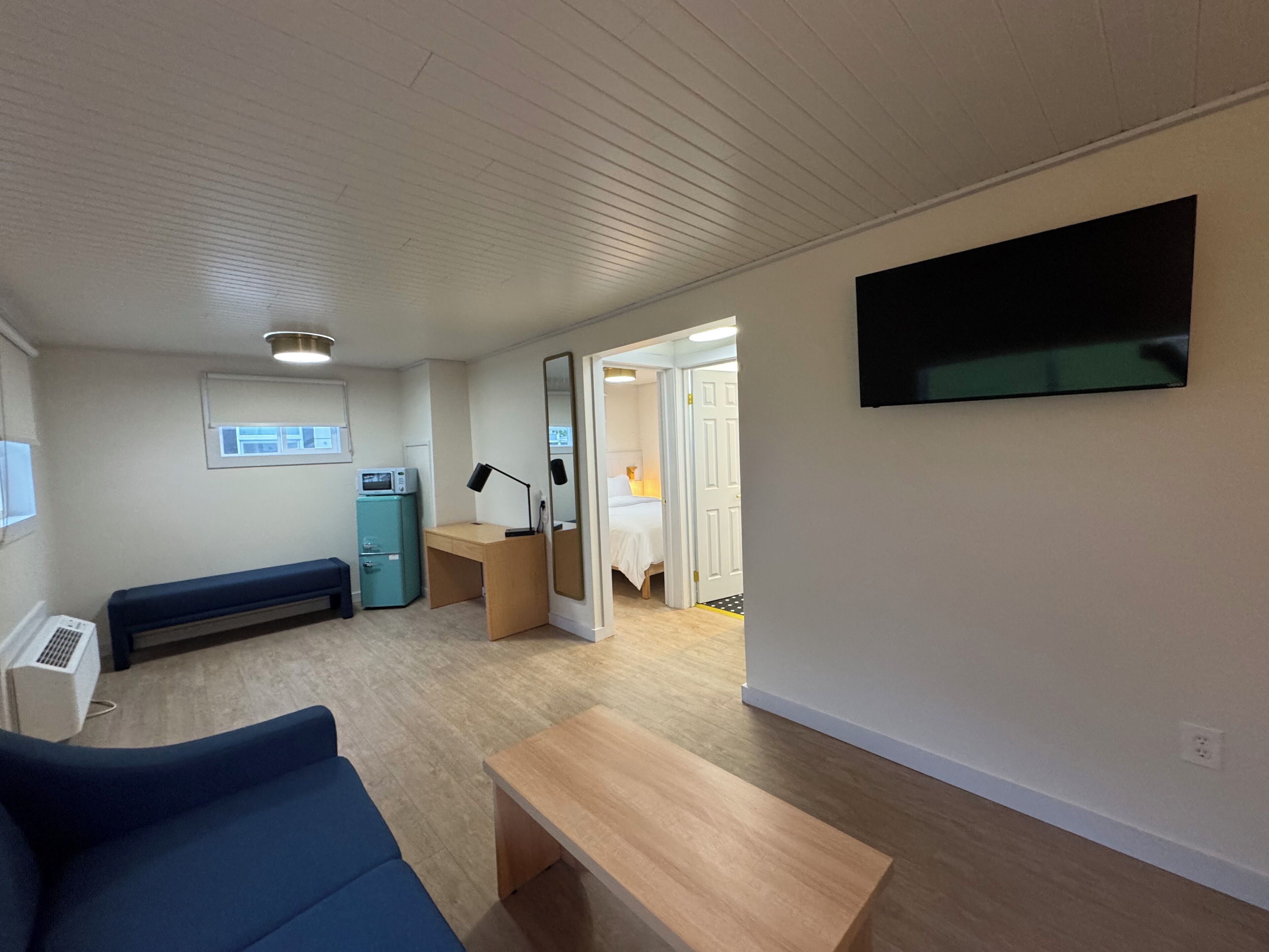 A compact living space with a sofa, a small desk, a mirror, a TV, and a kitchenette area.