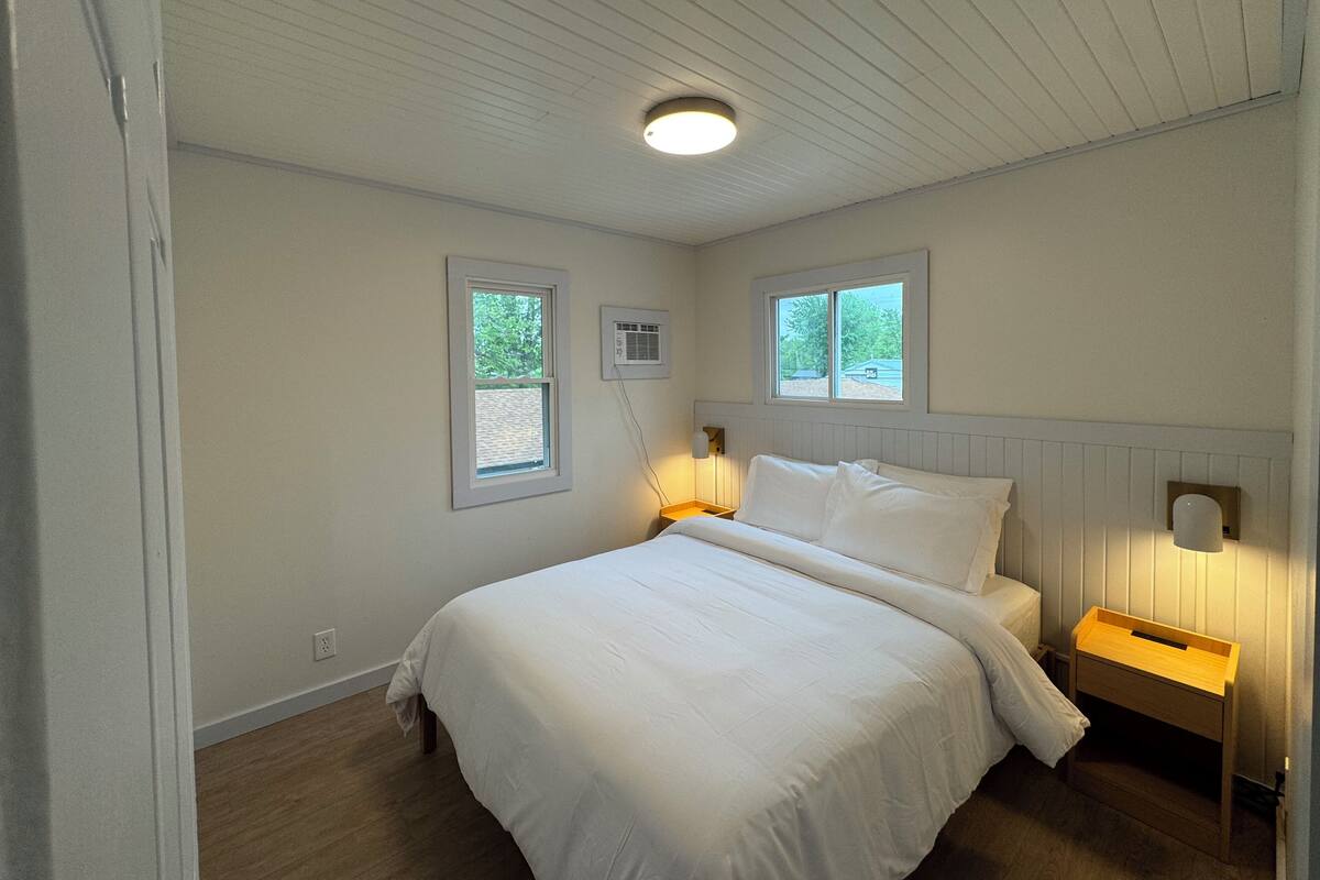 A bedroom with a bed, two windows, a ceiling light, a wall-mounted air conditioner, and a nightstand with a lamp.