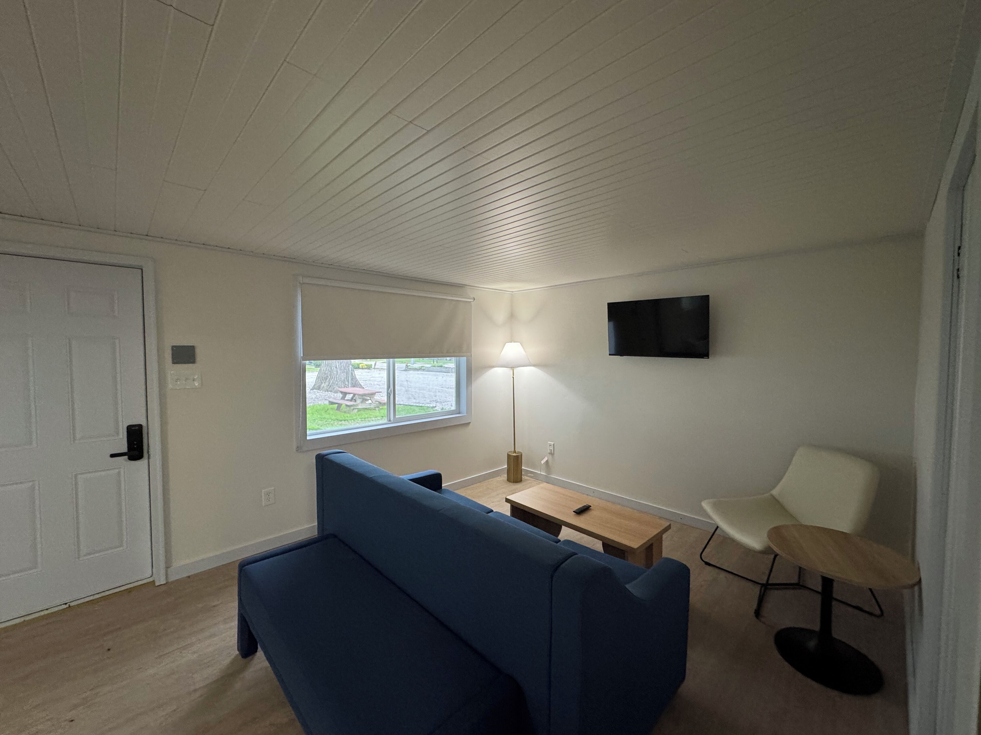 A room with a blue sofa, a small wooden table, a white chair, a lamp, and a TV mounted on the wall.