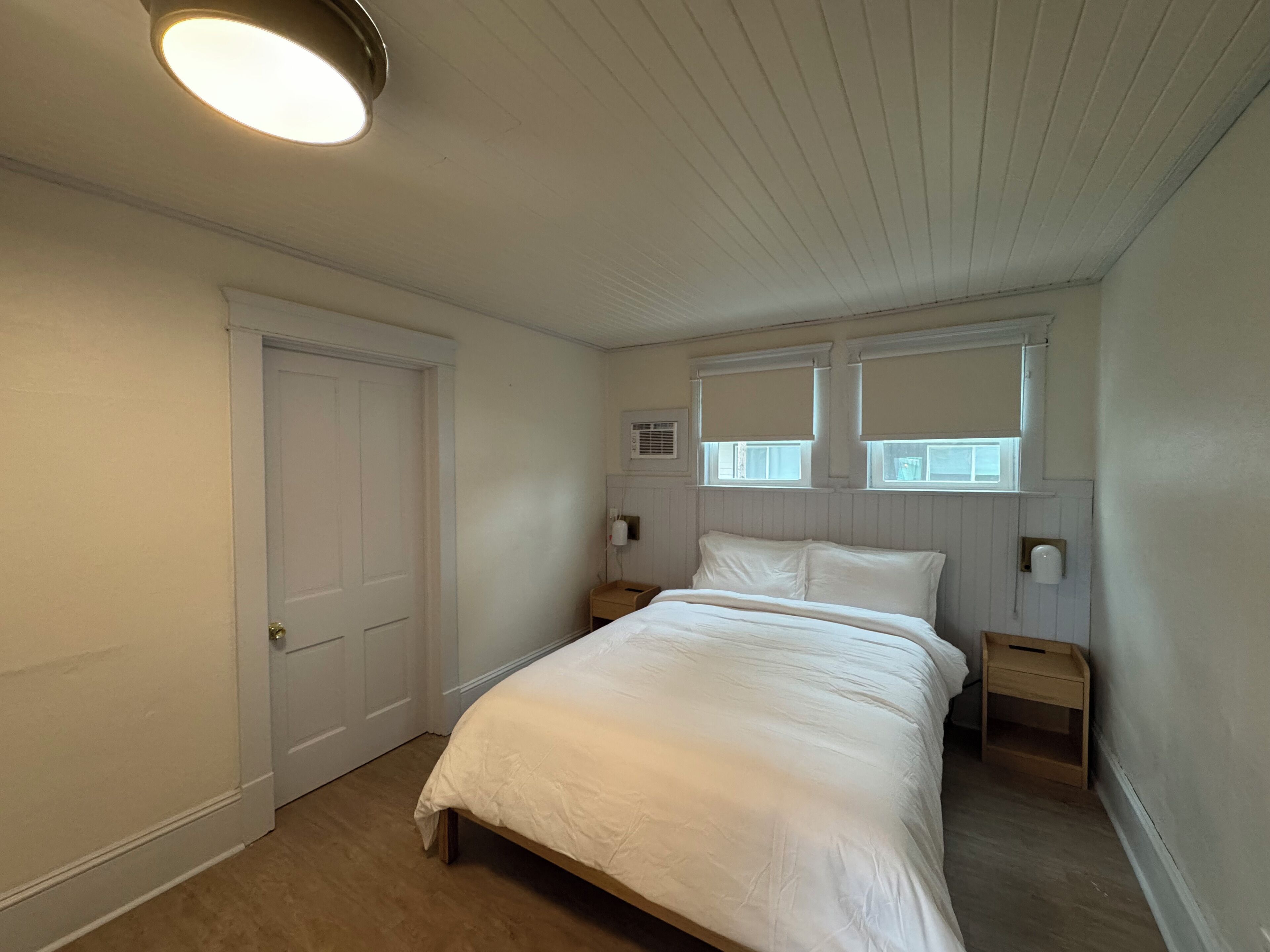 A bedroom with a bed, a door, a window with blinds, a nightstand, and a wall-mounted light fixture.