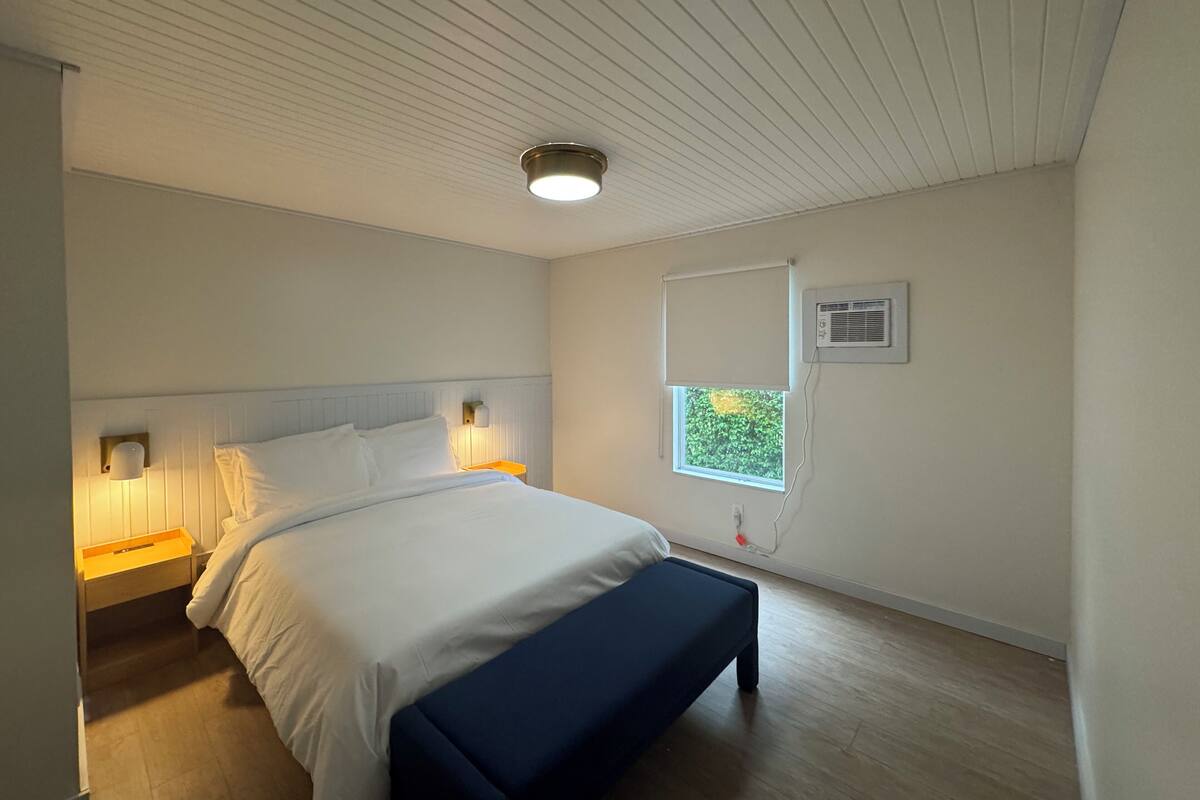 A bedroom with a bed, bedside lamps, a bench, a window with a view, and a ceiling light.