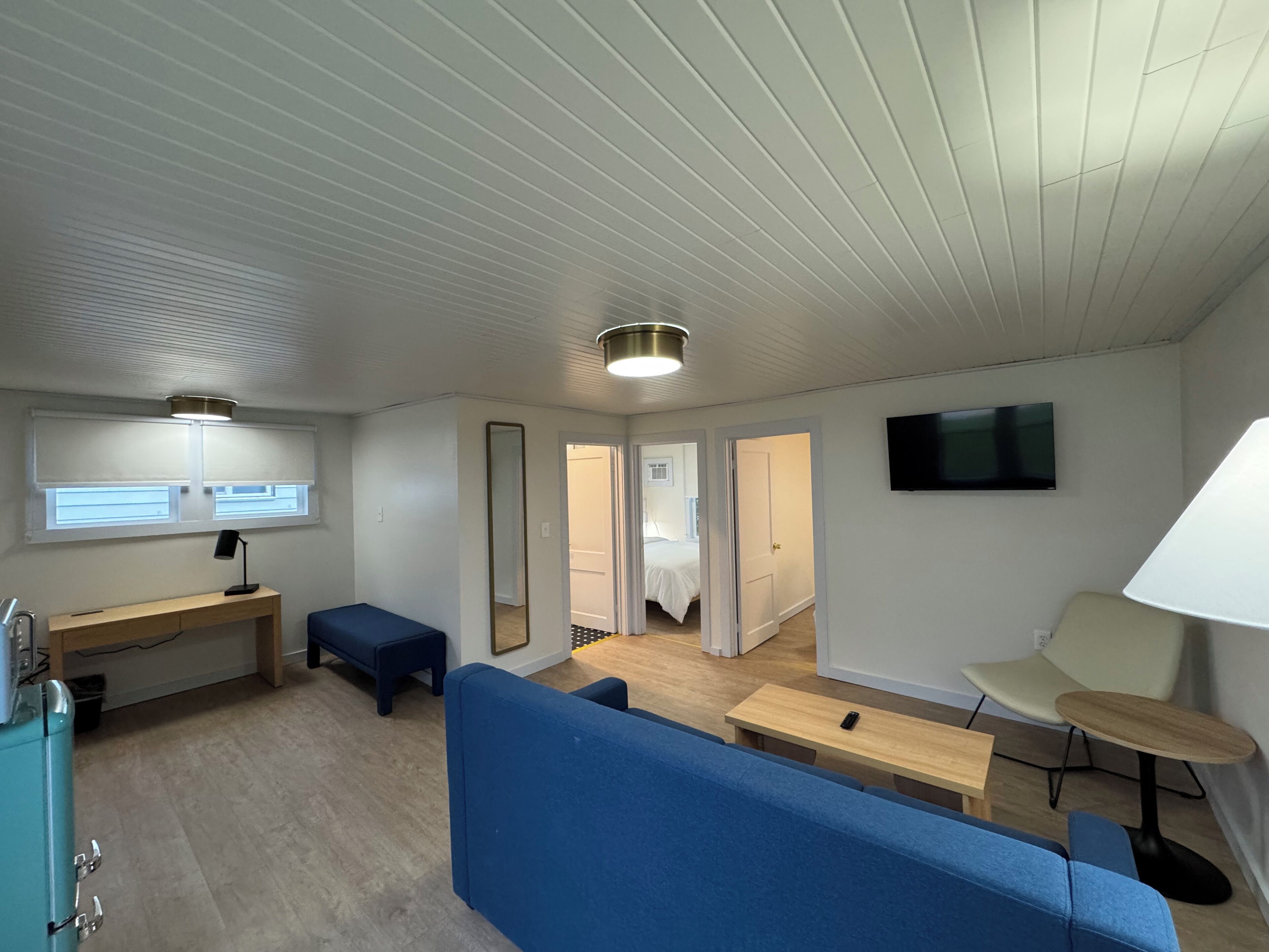 A compact room with a desk, a blue sofa, a small table, a TV mounted on the wall, and a mirror.