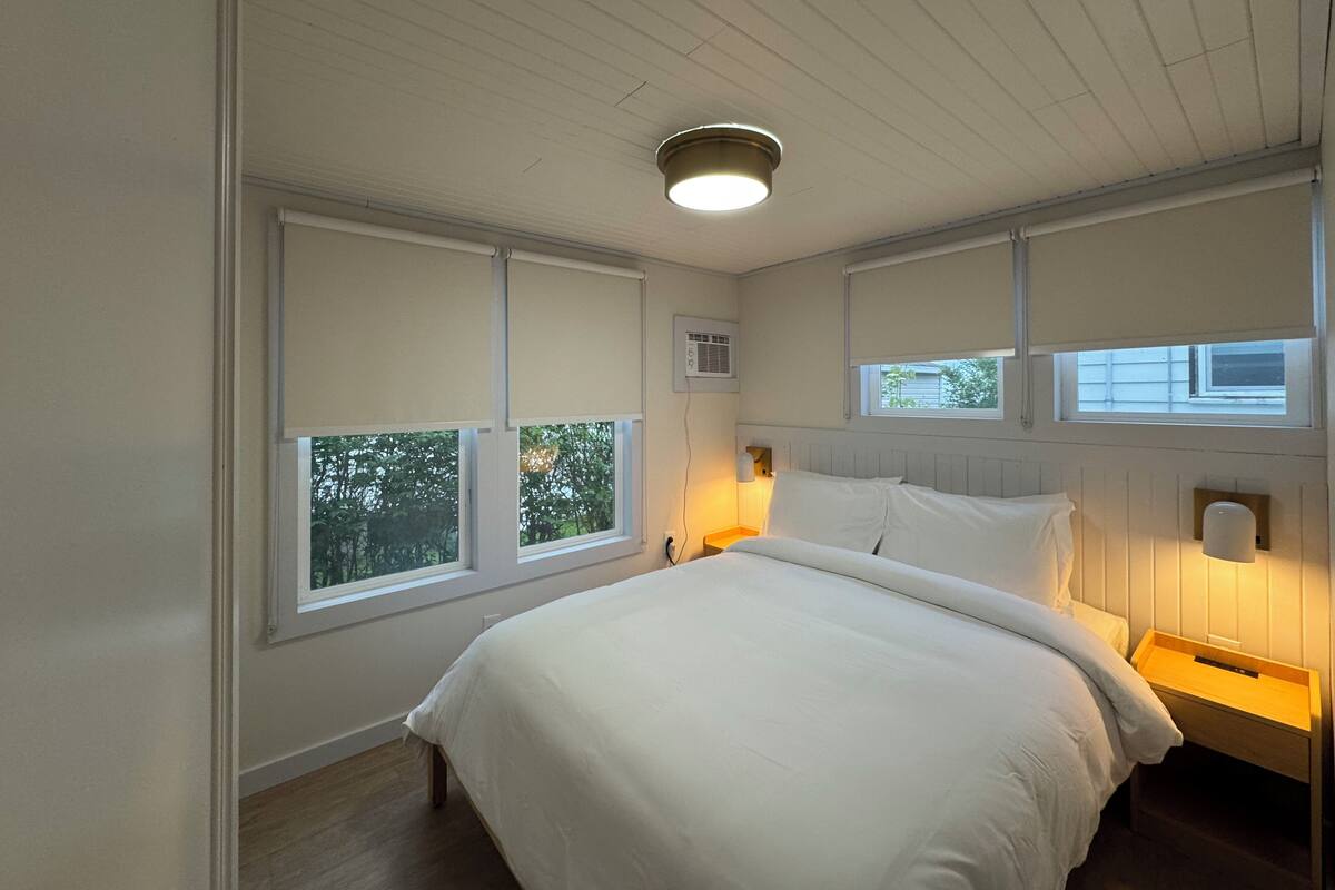 A bedroom with a large bed, two bedside tables, a ceiling light, and windows with blinds.