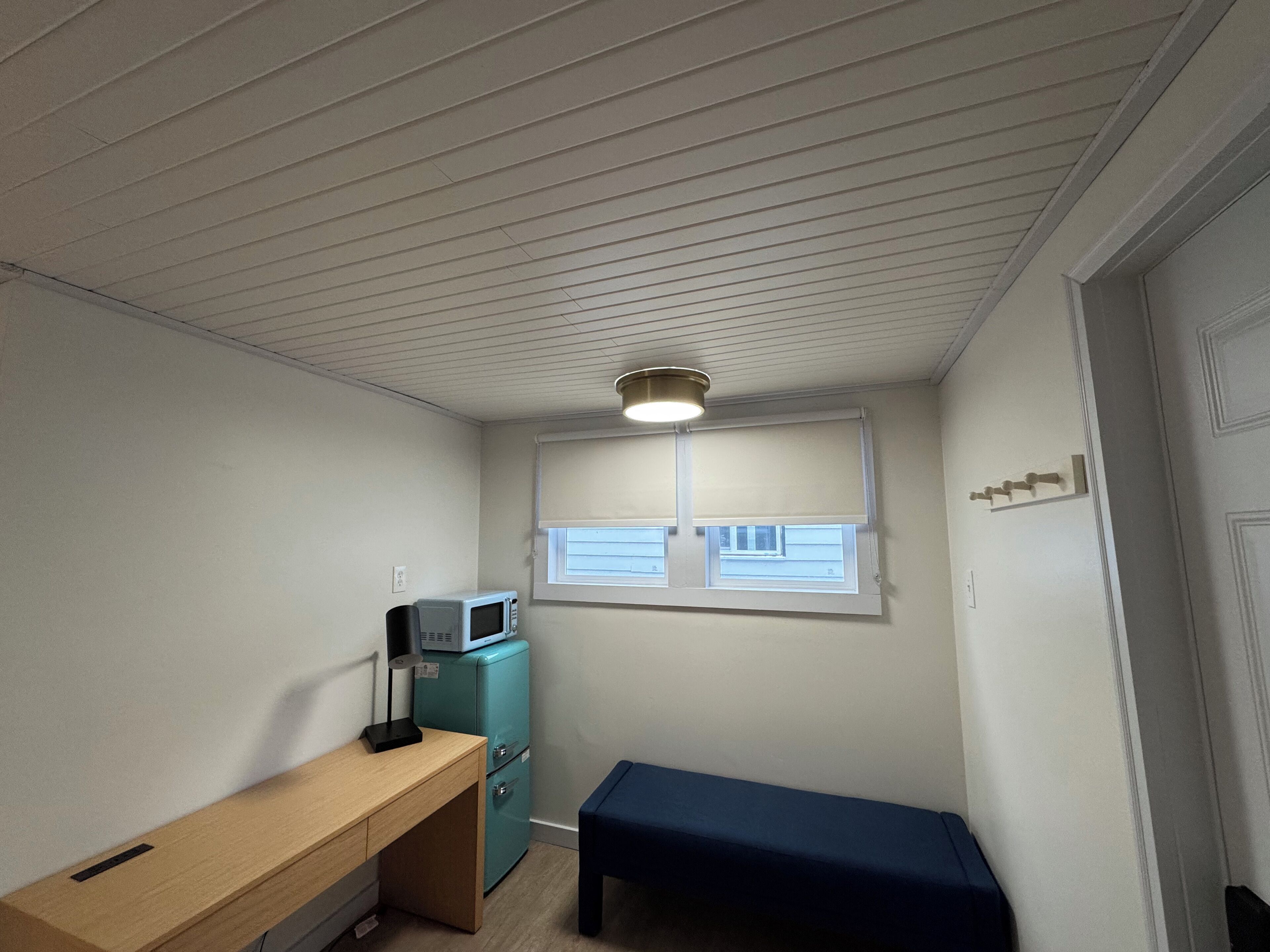 Um quarto pequeno com escrivaninha de madeira, um banco azul, geladeira verde-água e micro-ondas, além de uma janela com persianas.