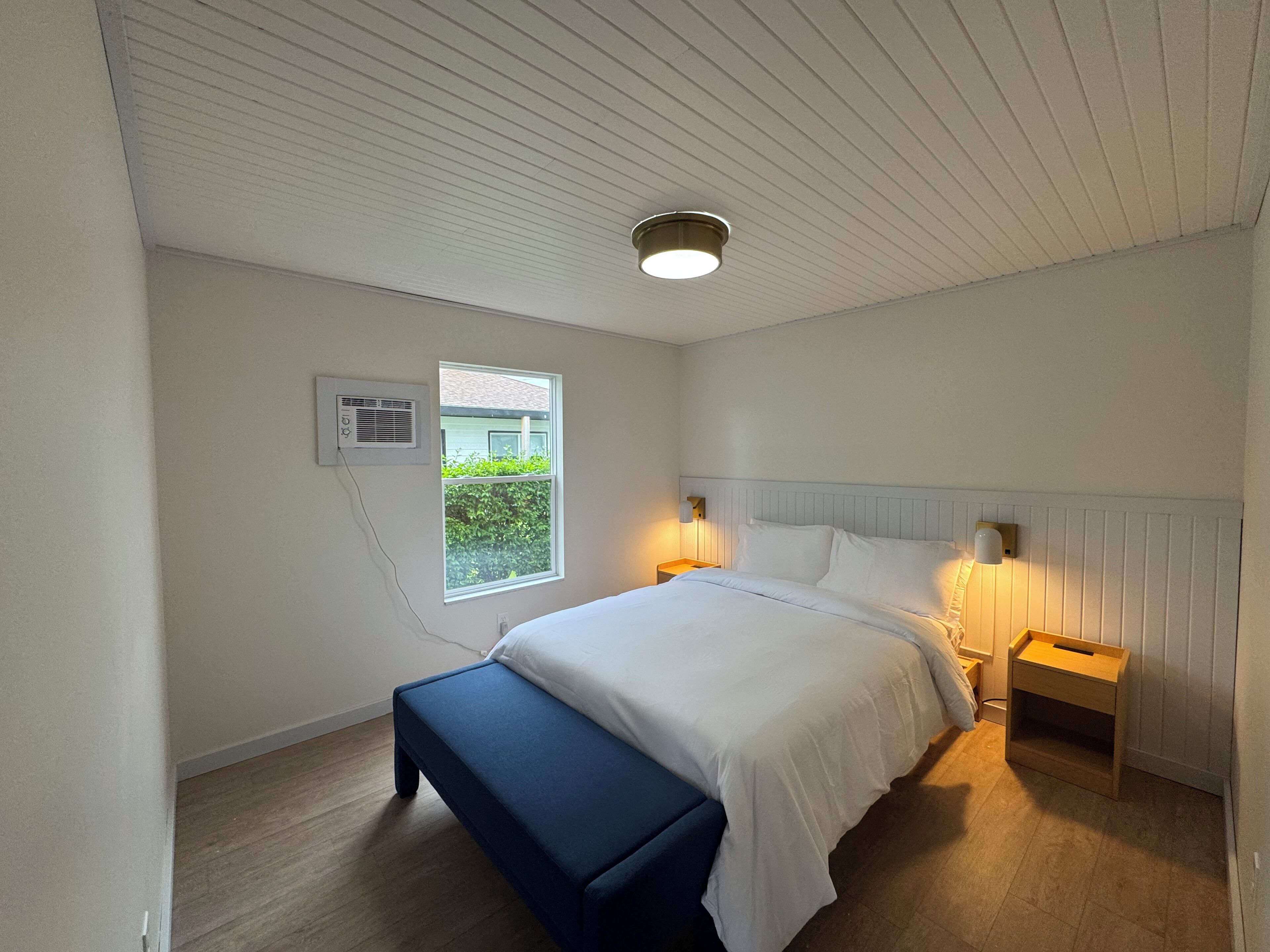 A bedroom with a bed, bedside tables, a bench, a window, and an air conditioning unit.