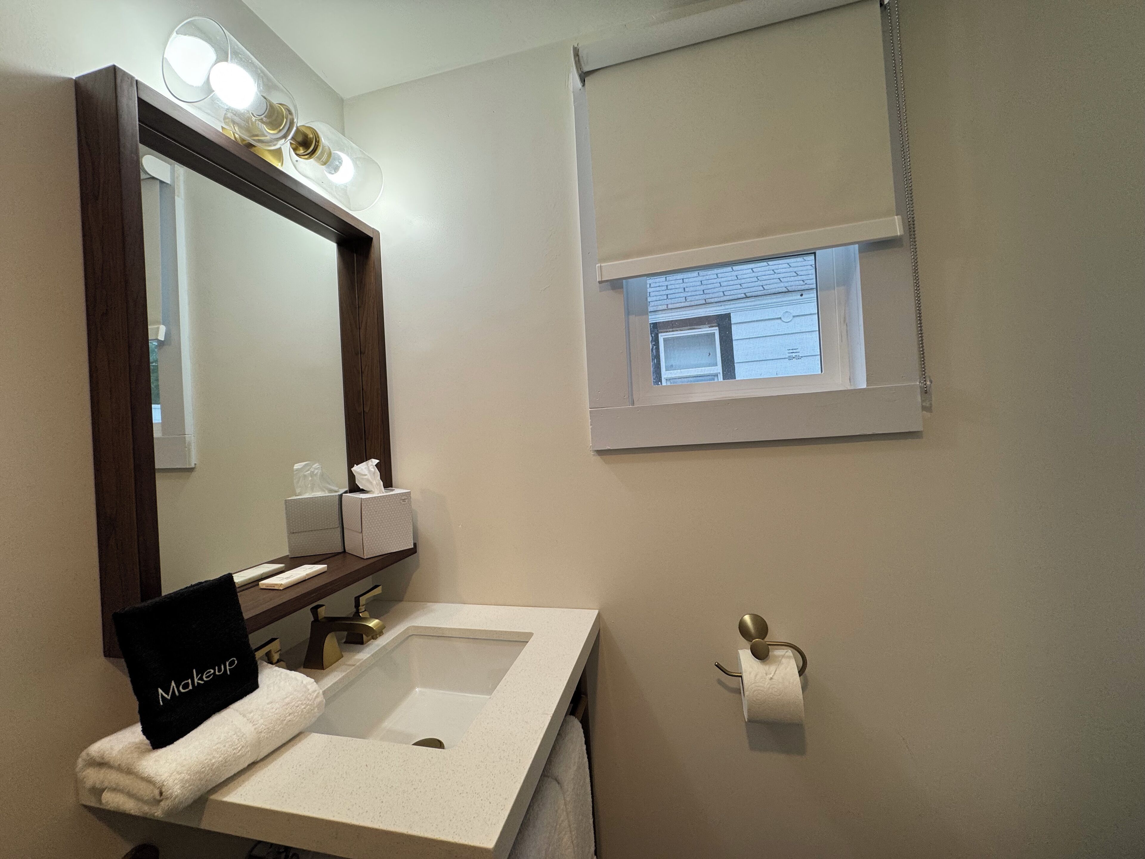 A bathroom with a vanity, a mirror, a sink, a towel, a tissue dispenser, and a window with a view of the outside.