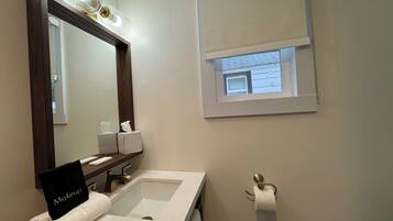 A bathroom with a vanity, a mirror, a sink, a towel, a tissue dispenser, and a window with a view of the outside.