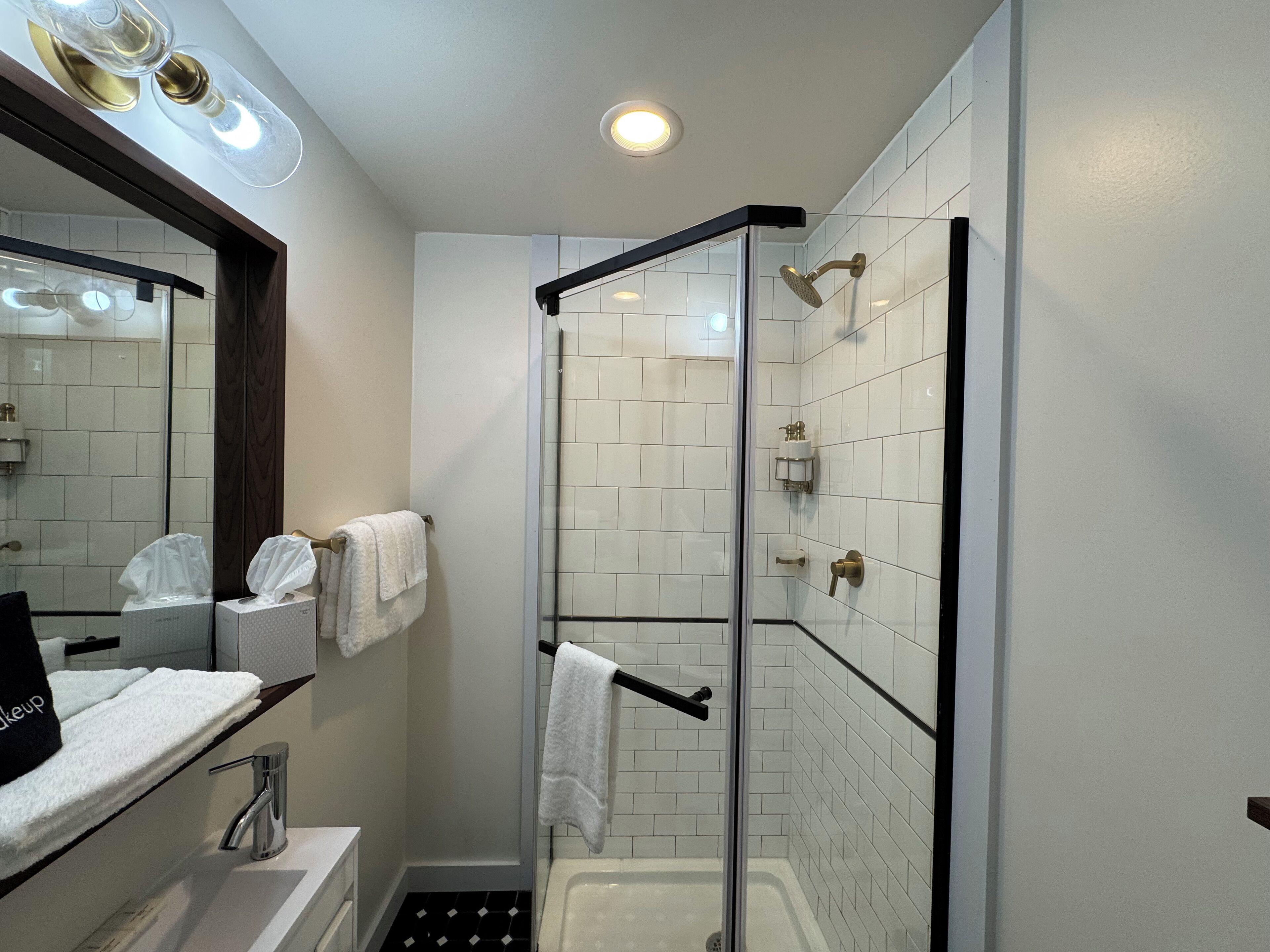 A modern bathroom with a glass-enclosed shower, white tiled walls, a built-in sink, and a mirror with lights above.