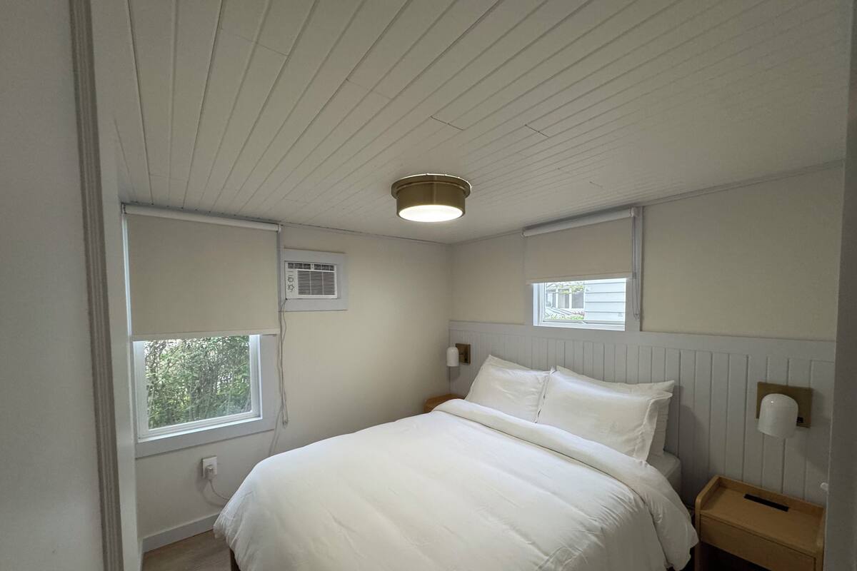 A bedroom with a bed, a bedside table, a window, and a ceiling light.