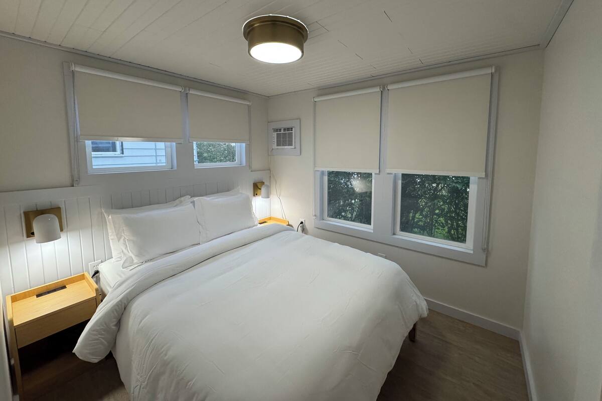 A bedroom with a large bed, a nightstand, a wall-mounted lamp, and two windows with blinds.