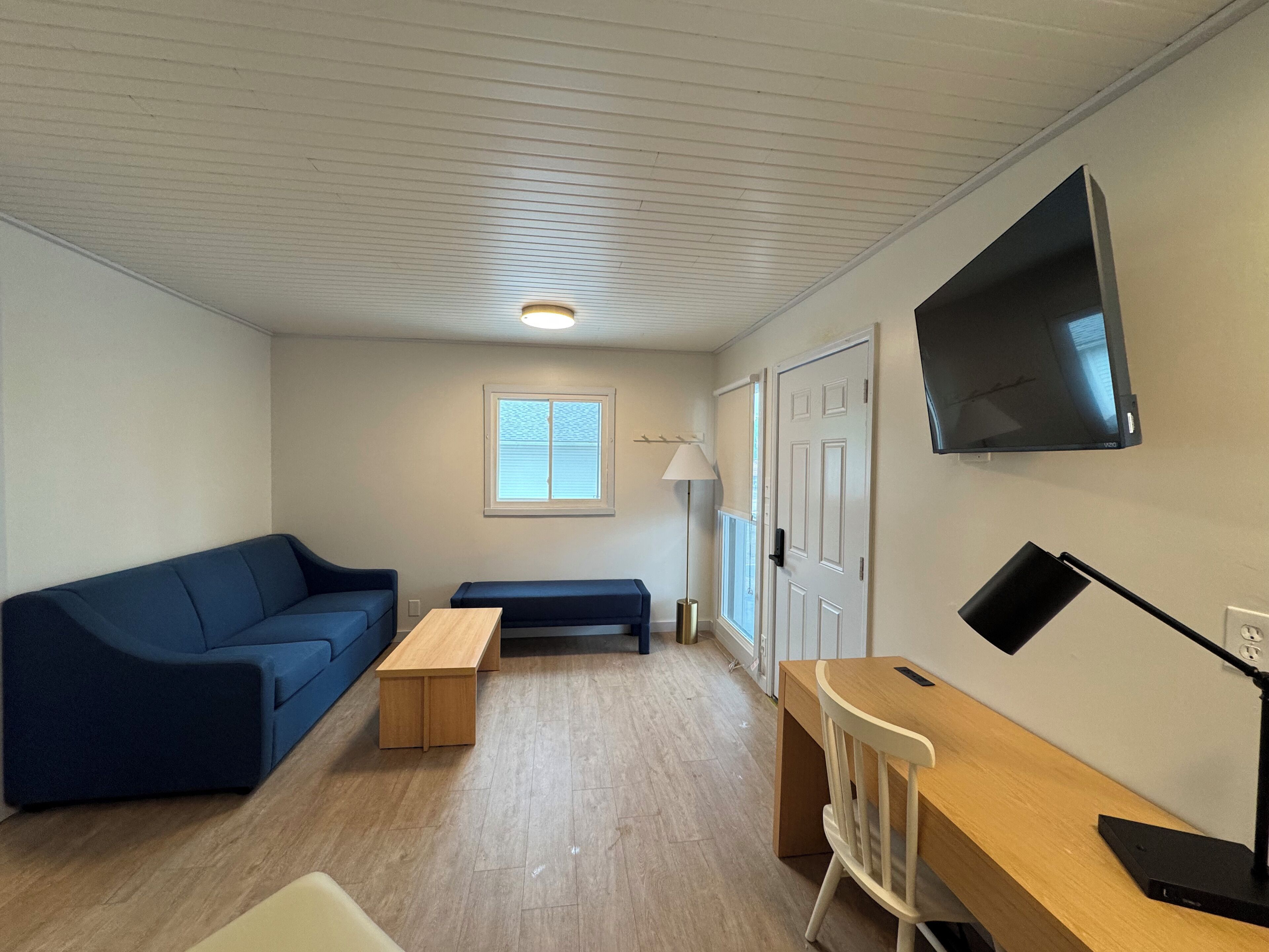 A room with a blue sofa, a wooden table, a white chair, a TV mounted on the wall, and a window with blinds.