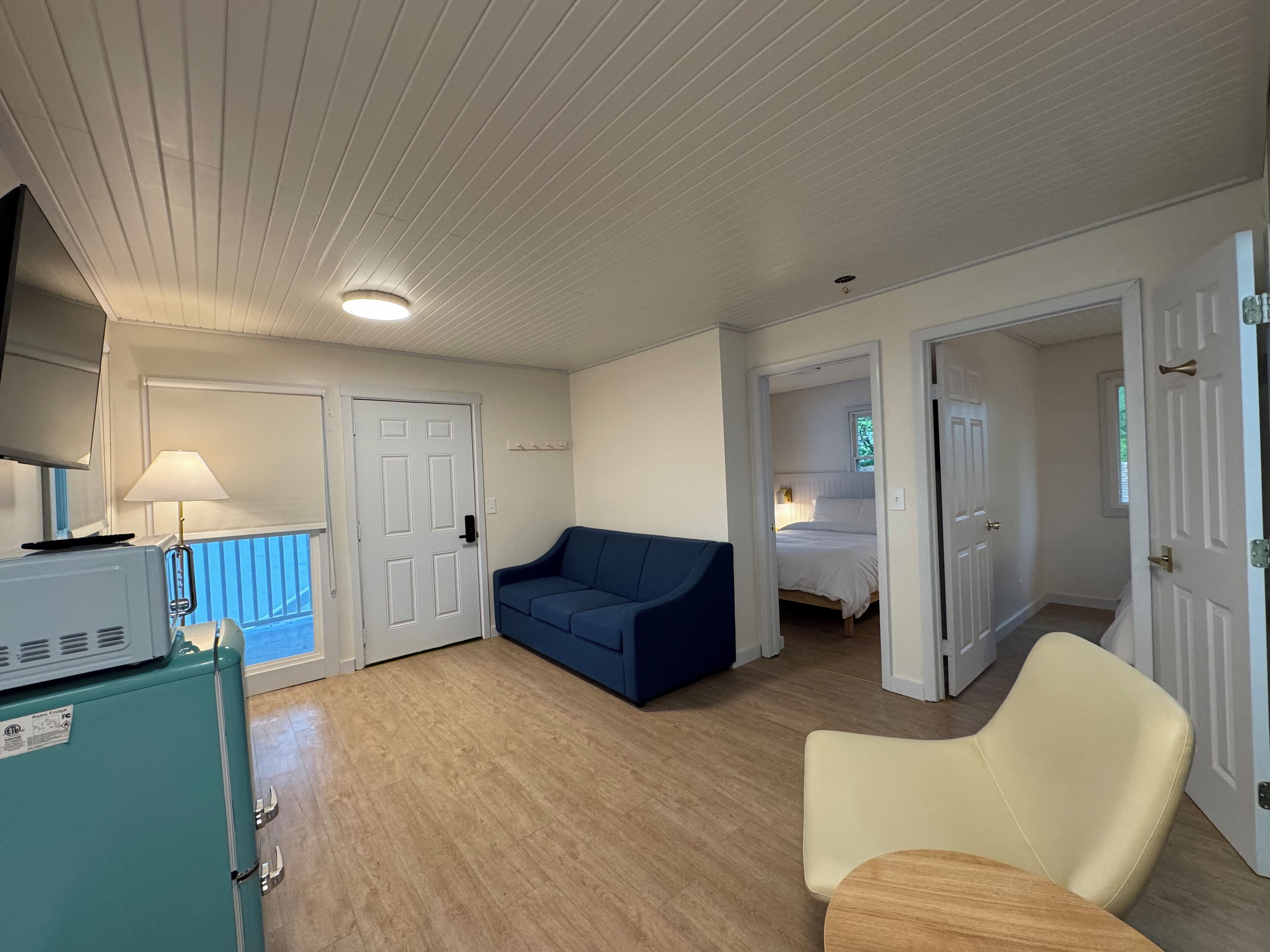 Una habitación con un sofá azul, una silla blanca, una cama en la habitación contigua y una kitchenette con un refrigerador.