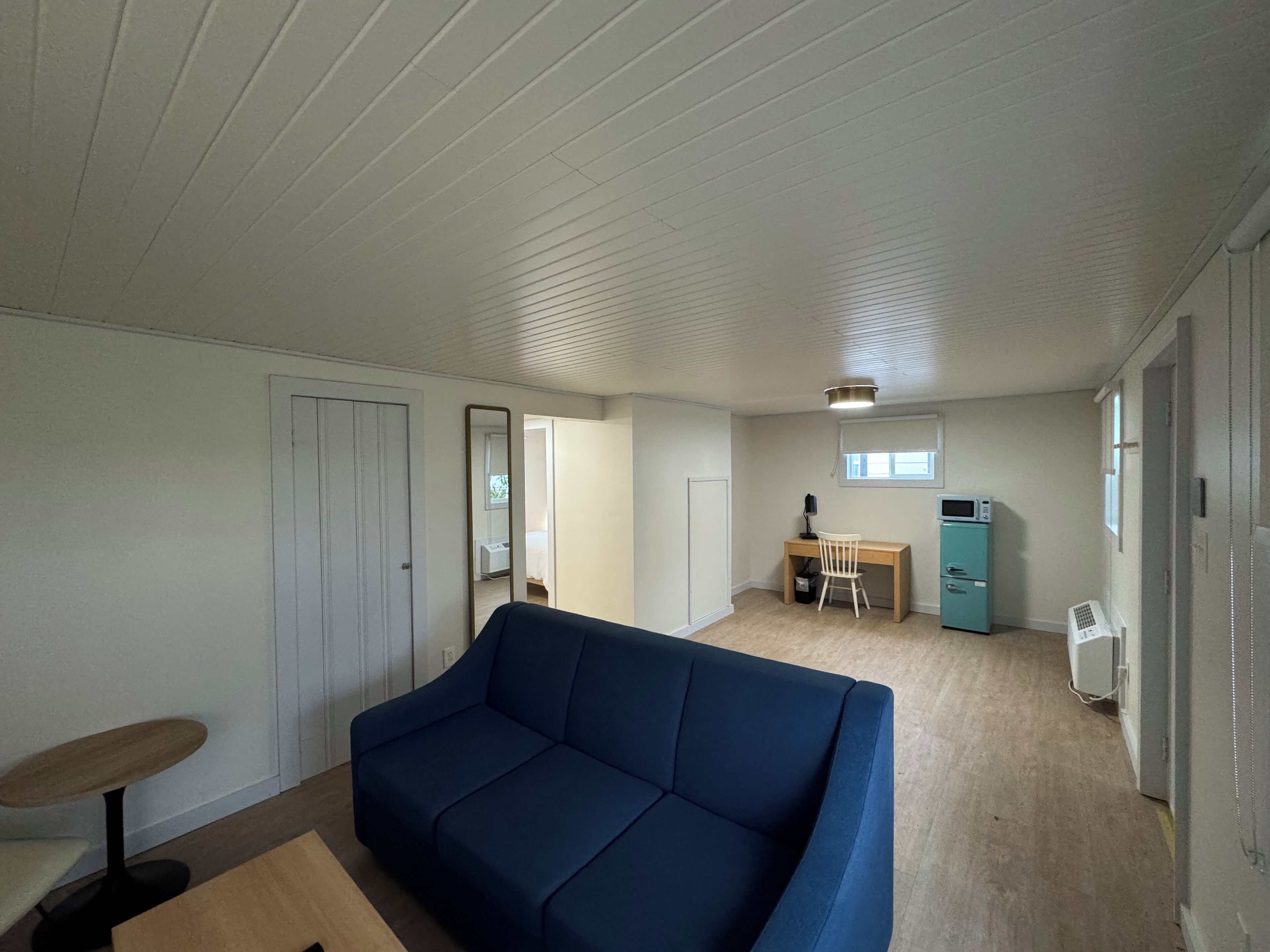 A room with a blue sofa, a desk with a chair, a small kitchenette, and a door leading to another room.