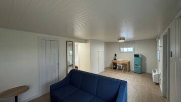 A room with a blue sofa, a desk with a chair, a small kitchenette, and a door leading to another room.