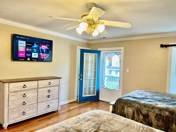 Une chambre dotée d’un ventilateur de plafond, d’un téléviseur fixé au mur qui diffuse différents services de streaming, d’une commode en bois, d’une porte avec un cadre bleu et d’une fenêtre avec des stores.