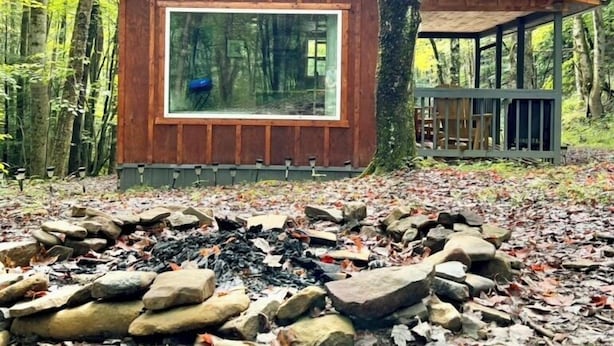 Een blokhut met een groot raam, omringd door bomen en een vuurkuil.