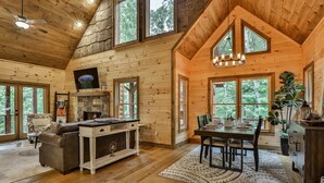 A log cabin interior with a fireplace, dining area, and large windows. - Happy Hideaway | Mineral Bluff, Ga (Mineral Bluff)